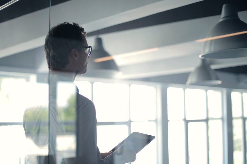 A businessman holding a tablet in an office, looking thoughtfully through a glass wall.
